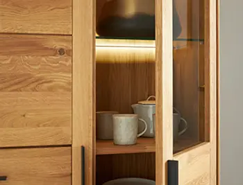 Detailaufnahme eines hochwertigen Eichenholz-Sideboards mit Glastür und integrierter LED-Beleuchtung – stilvolle Präsentation von Keramikgeschirr in einem modernen Wohnzimmer.