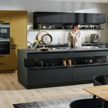 Schwarze Küche mit Insel und Küchenzeile an der Wand. In der Küche steht eine Frau und liest in einem Buch.