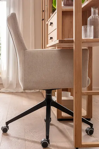Seitenansicht von Armlehnstuhl in beigem Stoff mit schwarzem Gestell und Rollen an einem Schreibtisch.