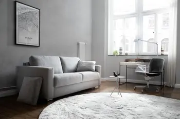 Hellgraues Zweisitzer Sofa mit grauem Kissen in einem hellen Raum mit großen Fenstern, vor denen  Vorhänge hängen.