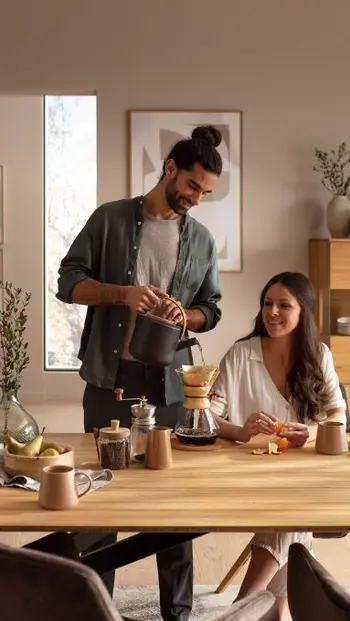 Mann steht neben einer sitzenden Frau am Esstisch und brüht Kaffee auf.