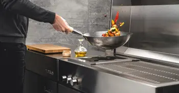 Mann schwingt Wokpfanne mit verschiedenem Gemuese ueber Heizflaeche einer Outdoorkueche. 