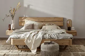 Massivholzbett mit hellen Kissen und Decke und Blumen auf dem Nachttisch daneben.