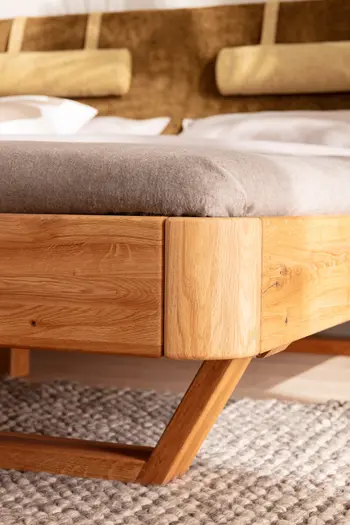 Abgerundete Ecke eines Bettes aus der Nähe, das Bett hat einen Kufenfuß und ist aus Massivholz.