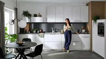 Moderne Küche in Weiß mit dunkler Arbeitsplatte und minimalistischer Ausstattung – eine Frau lehnt entspannt an der Küchenzeile und genießt ihr Getränk. Himmlisch Wohnen Blanco Choice Icona Unit