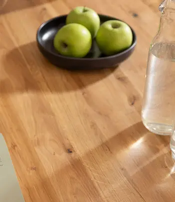 Tischplatte aus massiver Wildeiche, dekoriert mit einer Schale grüner Äpfel und einer Glasflasche.