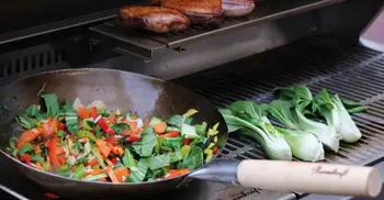 Verschiedenes Gemüse in einer Wokpfanne auf der Grillflaeche einer Outdoorkueche. 