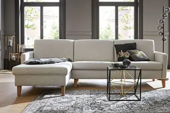 Hellgraue Eckcouch mit Holzfüßen auf dem dunkle Kissen und ein schwarzes Fell liegen in einem hellen Raum.