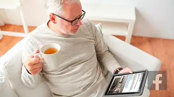 Ein Mann liest auf der Couch einen Facebookeintrag auf seinem Tablet und trinkt dabei Tee.&nbsp;