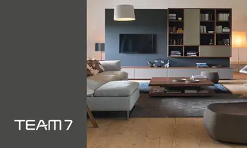 Ein Wohnzimmer mit Sofa, Wohnwand und Bestelltisch der Marke Team7, auf der linken Seite befindet sich das Team7-Logo&nbsp;auf grauem Hintergrund.