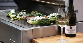 Grillflaeche einer Outdoorkueche mit verschiedenem Gemuese und Fleisch neben einer Weinflasche mit Glas. 