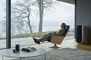 Ein Mann entspannt sich in einem Stressless Sam Sessel mit Holzseiten in der Farbe Paloma Rock und einer runden Basis aus Eiche. Der Raum bietet durch große Fenster einen Blick auf eine ruhige Landschaft mit Bäumen und Wasser.