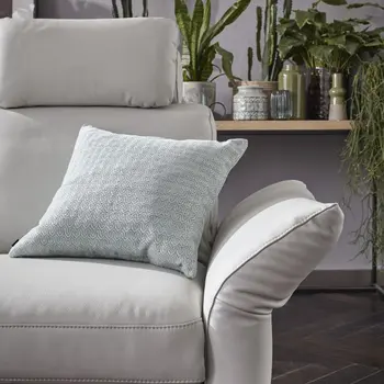 Hellgraues Ledersofa mit Kopfstütze und weggeklappter Armlehne, das mit einem hellen Kissen mit Muster dekoriert ist, im Hintergrund sind viele Pflanzen sichtbar.