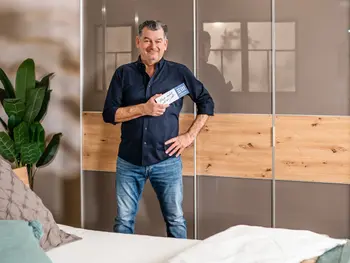 Der Markenbotschafter von der Europa Möbel Collection, Mark Kühler, steht vor einem großen Kleiderschrank und hält einen Flyer in die Kamera.  