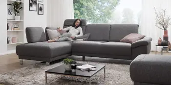 Frau entspannt auf dunkelgrauem Stoff-Ecksofa in hellem Wohnzimmer. 