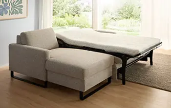 Modernes Schlafsofa mit beigem Stoffbezug und schwarzem Metallgestell im ausgezogenen Zustand – platzsparende Schlafmöglichkeit im lichtdurchfluteten Wohnzimmer mit Ausblick ins Grüne.