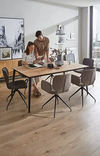 Modernes Esszimmer mit großem Esstisch aus Holz und sechs gepolsterten Schalenstühlen mit Metallgestell – ein Paar bespricht Unterlagen in heller, stilvoll eingerichteter Wohnatmosphäre mit urbanem Ausblick.