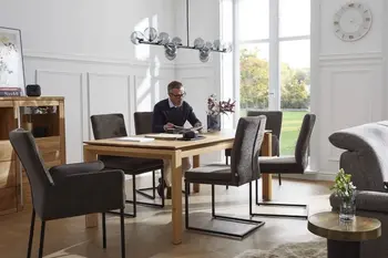 Mehrere graue Freischwinger mit schwarzem Metallgestell und zwei graue Armlehnstühle um einen Holz Esstisch, auf einem der Stühle sitzt ein Mann und liest.