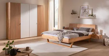 Helles gemütliches Schlafzimmer mit Kleiderschrank in hellem Holz mit Lamellenoptik und weißen Elementen an den Türen sowie ein dazu passendes Bett mit Lamellenoptik und weißen Elementen.