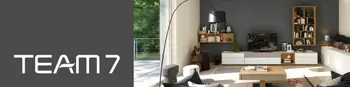 Modische Wohnwand mit weißen Fronten und Detail in Holz. Davor ein passender TV-Tisch. Links neben dem Bild, das Team7 Logo auf grauem Hintergrund.&nbsp;