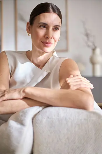 Elegant gekleidete Frau sitzt entspannt auf beigem Sofa und stützt sich auf Armlehne.