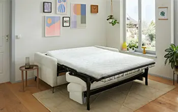 Weißes Schlafsofa mit schwarzem Metallgestell im ausgezogenen Zustand – multifunktionales Möbelstück im gemütlich eingerichteten Raum mit Wandbildern, Pflanzen und natürlichem Tageslicht.