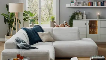 Hellgraues Ecksofa aus Cord, dekoriert mit mehreren Kissen, hinter der Rückenlehne sieht man ein Kind, das mit Handpuppen spielt.