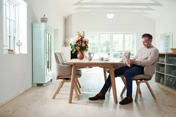 Esszimmer mit Stressless Laurel Stühlen in Calido Light Beige und Eichenholzbeinen, um einen Holztisch. Ein Mann liest eine Zeitung in einem lichtdurchfluteten Raum mit Blumen und klassischem Dekor.