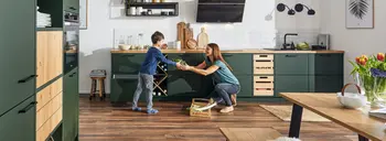 Frau mit Kind in der Küche mit großer dunkelgrüner Küchenzeile mit Holzarbeitsplatte.&nbsp;&nbsp;