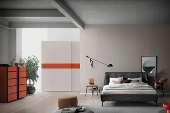 Großer weißer Schwebetürenschrank, der mittig einen orangen Akzent hat in einem hellen Schlafzimmer mit grauem Polsterbett.