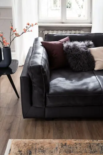 Linker Teil eines modernen schwarzen Ledersofas mit hoher dick gepolsterter Armlehne und Dekokissen.