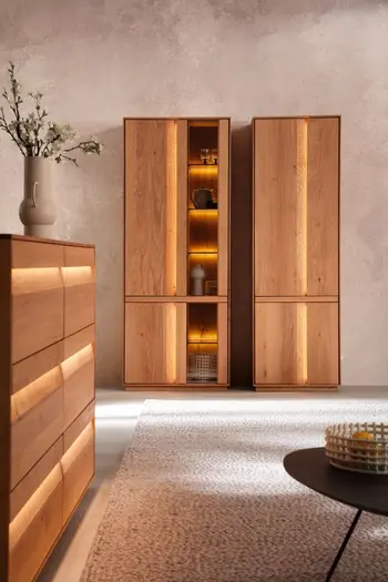 Große Vitrine aus Eichenholz mit Glasetür rechts neben einem Vollholz Schrank, seitlich davon steht ein Sideboard mit beleuchteten Griffen Fronten.