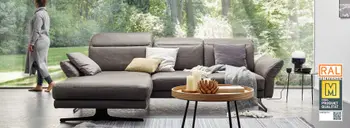 Ecksofa in grauem Leder vor einer großen Fensterfront mit Blick ins grüne.