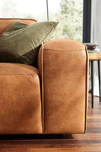 Abgerundete Armlehne eines Sofas aus Leder in Farbe Cognac, auf dem ein grünes Kissen liegt.