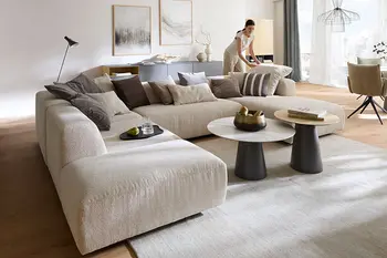 Frau in elegantem Wohnzimmer schüttelt Kissen auf beigem Ecksofa auf.