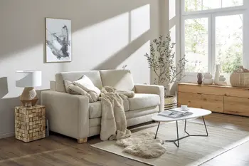 Beiges Sofa in einem lichtdurchfluteten Raum mit einem Beistelltisch daneben auf dem eine Lampe steht und einem modernen Couchtisch.