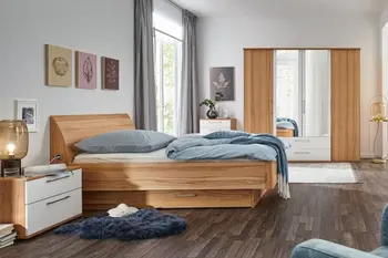 Helles Schlafzimmer in dem ein Massivholz Bett mit Stauraum, passender Nachttisch mit Schubladen mit weißer Front, sowie ein dazu passender Kleiderschrank mit Spiegel und weißen Akzenten steht.