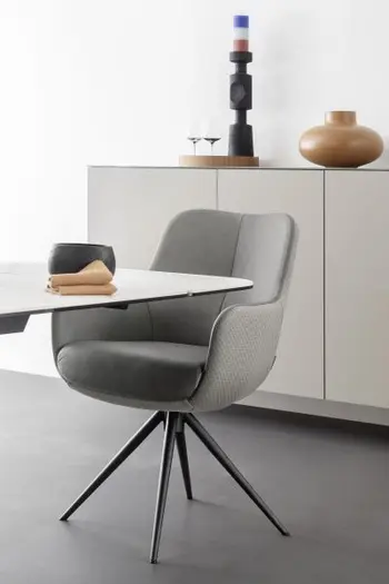 Ein grauer Armlehnstuhl, der innen mit Leder und außen mit Stoff bezogen ist und schwarzes Fußgestell hat und an einem Esstisch mit weißer Tischplatte steht.