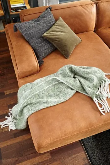 Longchair eines Sofas aus Leder in der Farbe Cognac, auf dem eine grüne Decke liegt und ein grünes und blaues Kissen.