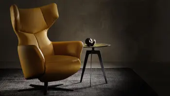 Moderner Designer Sessel mit Ohrenlehne, in gelbem Lederbezug mit schwarzem Sternfuß neben einem modernen dreibeinigen Beistelltisch mit schwarzem Gestell und goldener Metalltischplatte.