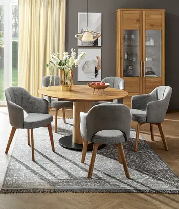 Runder Holztisch mit grauen Stühlen in Grau mit Holzfüßen. Im Hintergrund passende Vitrine.
