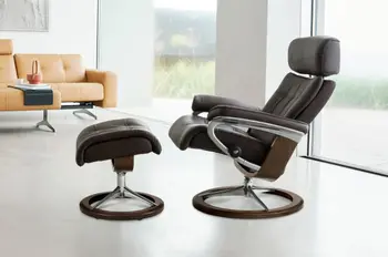 Stressless Erik Relaxsessel mit braunem Lederbezug und passendem Fußhocker, auf Signature-Untergestell aus Walnussholz und poliertem Metall, in einem modernen Wohnzimmer mit hellem Sofa im Hintergrund.