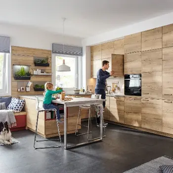 Ein Vater steht in einer Küche mit Holzfronten und bereitet Essen zu, während ein kleines Kind am integrierten Tisch der Küche spielt.&nbsp;