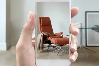 Eine Hand hält ein Smartphone, auf dessen Bildschirm ein Stressless Sessel in orangefarbenem Leder mit einem passenden Fußhocker und einer beigen Decke angezeigt wird. Im Hintergrund ein moderner Wohnraum mit minimalistischer Einrichtung.