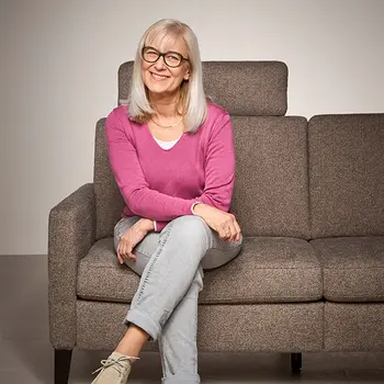Ältere Frau mit grauen Haaren und Brille sitzt entspannt auf einem ergonomischen Polstersofa in Braun – bequeme Couch mit verstellbarer Kopfstütze für modernes und komfortables Wohnen im Alter.