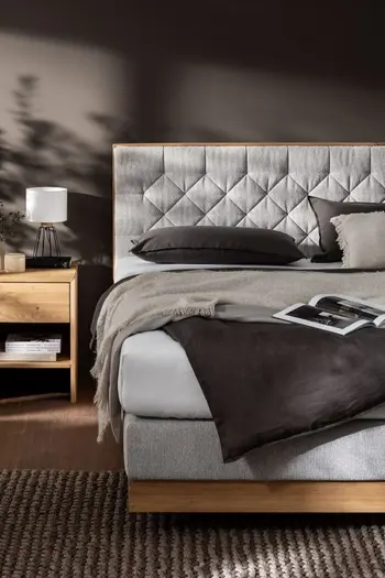 Hälfte eines modernen Boxspringbettes mit gestepptem grauem Kopfteil und Holzumrandung unten.