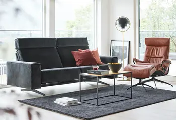 Modernes Wohnzimmer mit dunkelgrauem Sofa, braunem Relaxsessel und Couchtisch auf einem Teppich vor großen Fenstern.