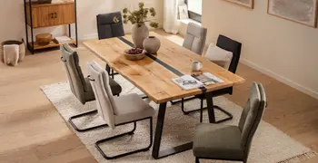 Modernes gemütliches Esszimmer mit einem großen Massivholzesstisch aus Wildeiche, der in der Mitte einen schmalen schwarzen Streifen aus Epoxidharz hat, um den Tisch stehen Freischwinger Stühle in grün, beige und grau.