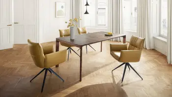 Designer Esstisch in Nussbaum mit filigranen Tischbeinen, um den Tisch stehen gelbe Armlehnstühle in einem hellen Raum.