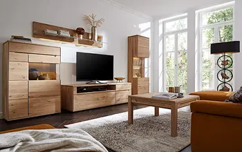 Modernes Wohnzimmer mit massiver Wohnwand aus Eiche – TV-Board, Vitrine, Regal und Couchtisch in natürlichem Holzdesign, kombiniert mit großen Fenstern, gemütlichem Sofa in Senfgelb und stilvollen Deko-Elementen für ein helles, wohnliches Ambiente.